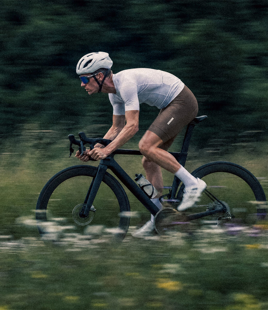 A cyclist in a white jersey and brown shorts rides a dark road bike on a blurred green path, wearing a white helmet and blue-tinted sunglasses.