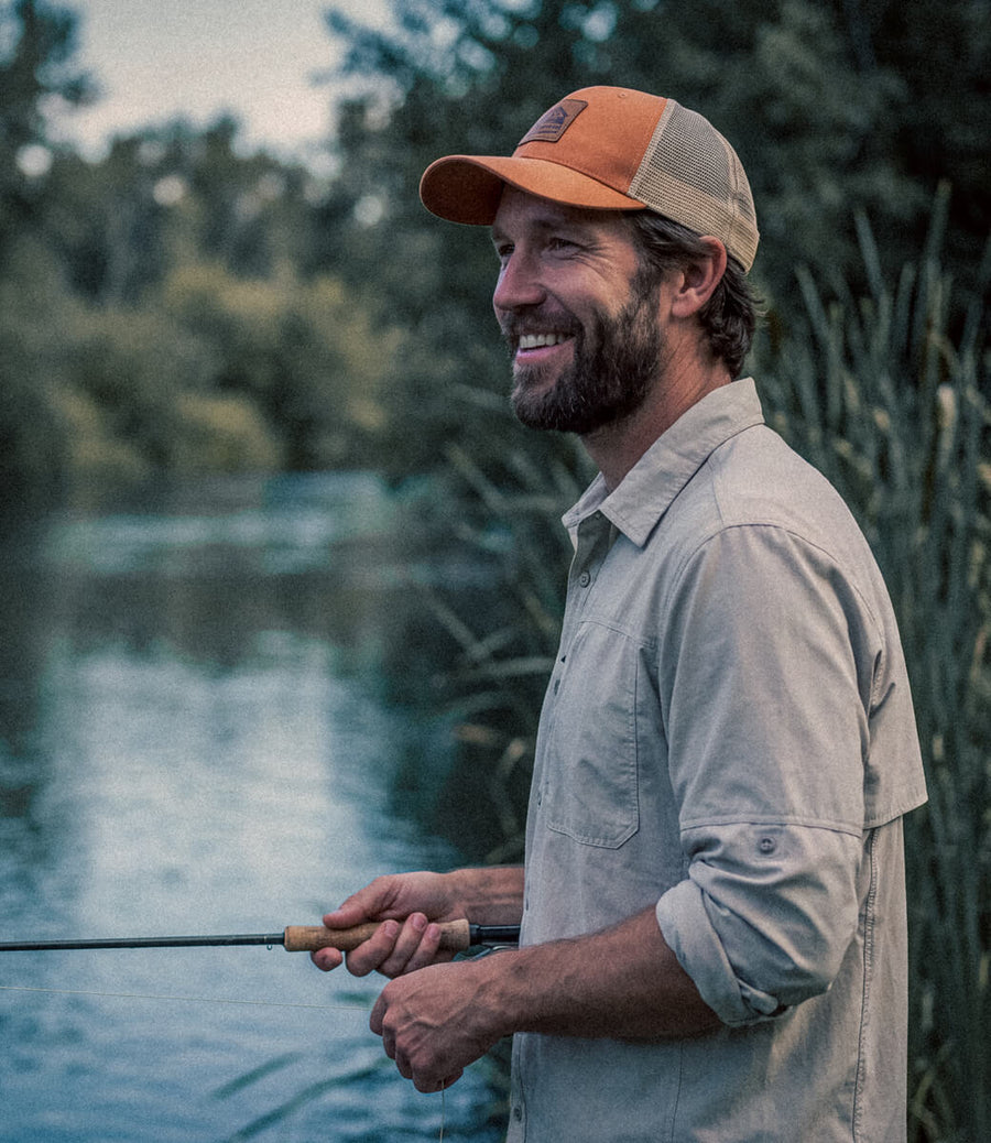A smiling man wearing a tan and orange trucker cap and a light beige button-up shirt stands by a river holding a fishing rod, with trees and reeds in the background.