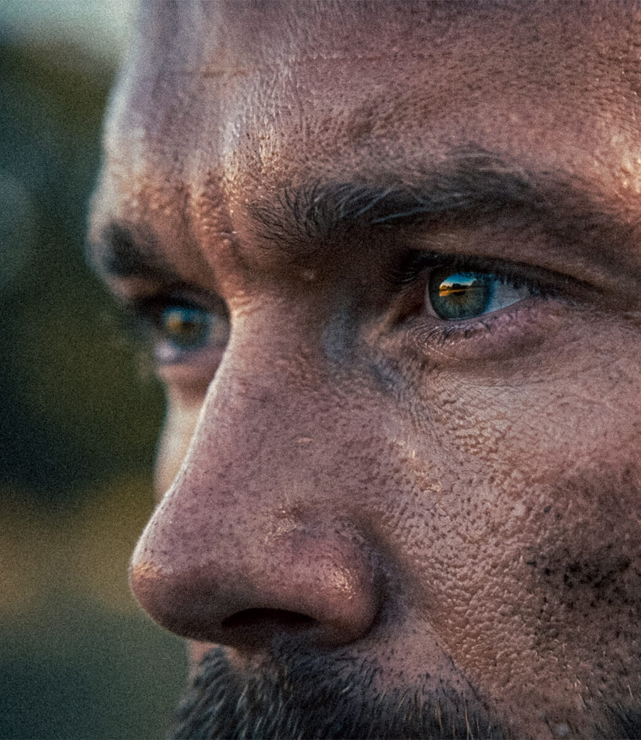 Extreme close-up of a man's face in profile, showing detailed skin texture, freckles, and blue eyes with a blurred outdoor background.