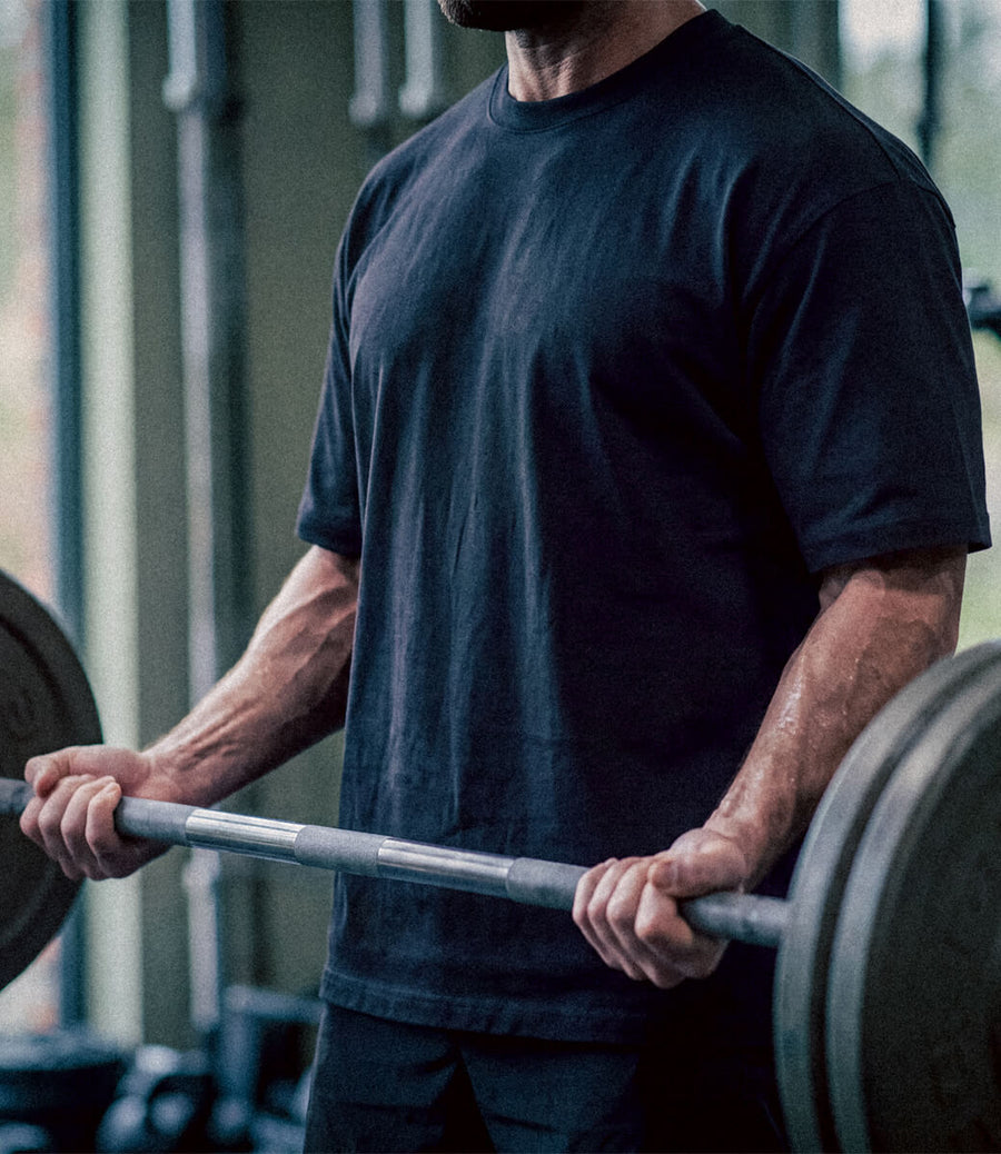 A man in a gym wearing a dark navy blue t-shirt and lifting a barbell with weight plates.
