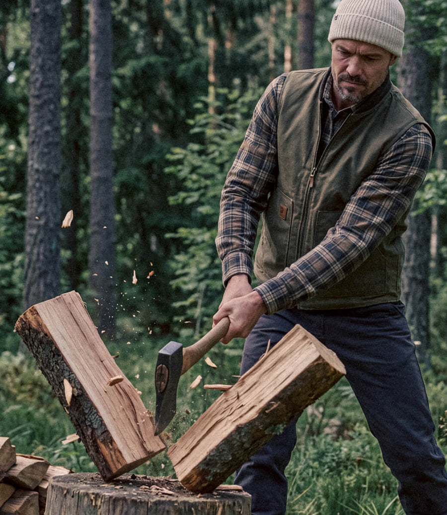 A man in a forest wearing a green quilted vest over a plaid shirt, swinging an axe to split wood on a stump.
