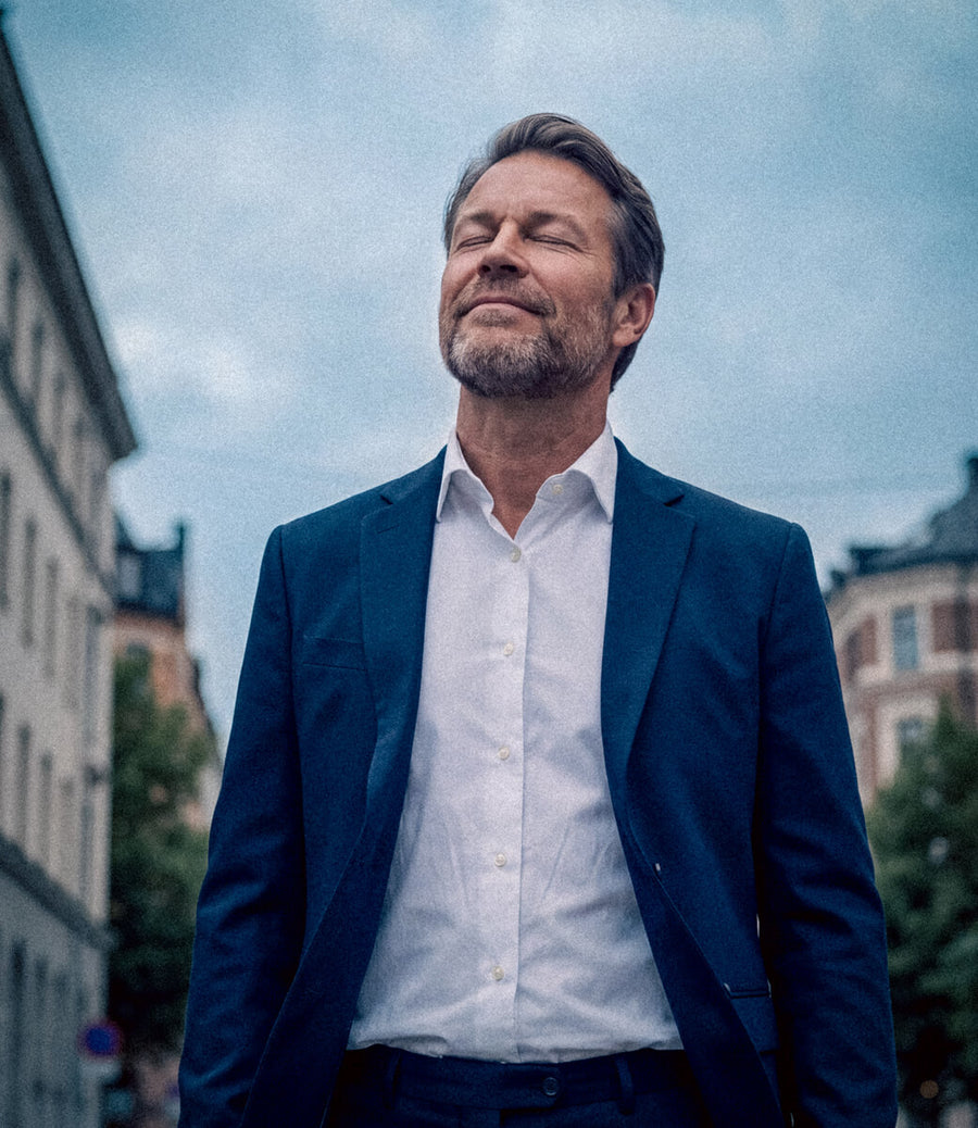 A man wearing a navy blue suit jacket and a white button-down shirt, standing outdoors with a cloudy sky and buildings in the background.