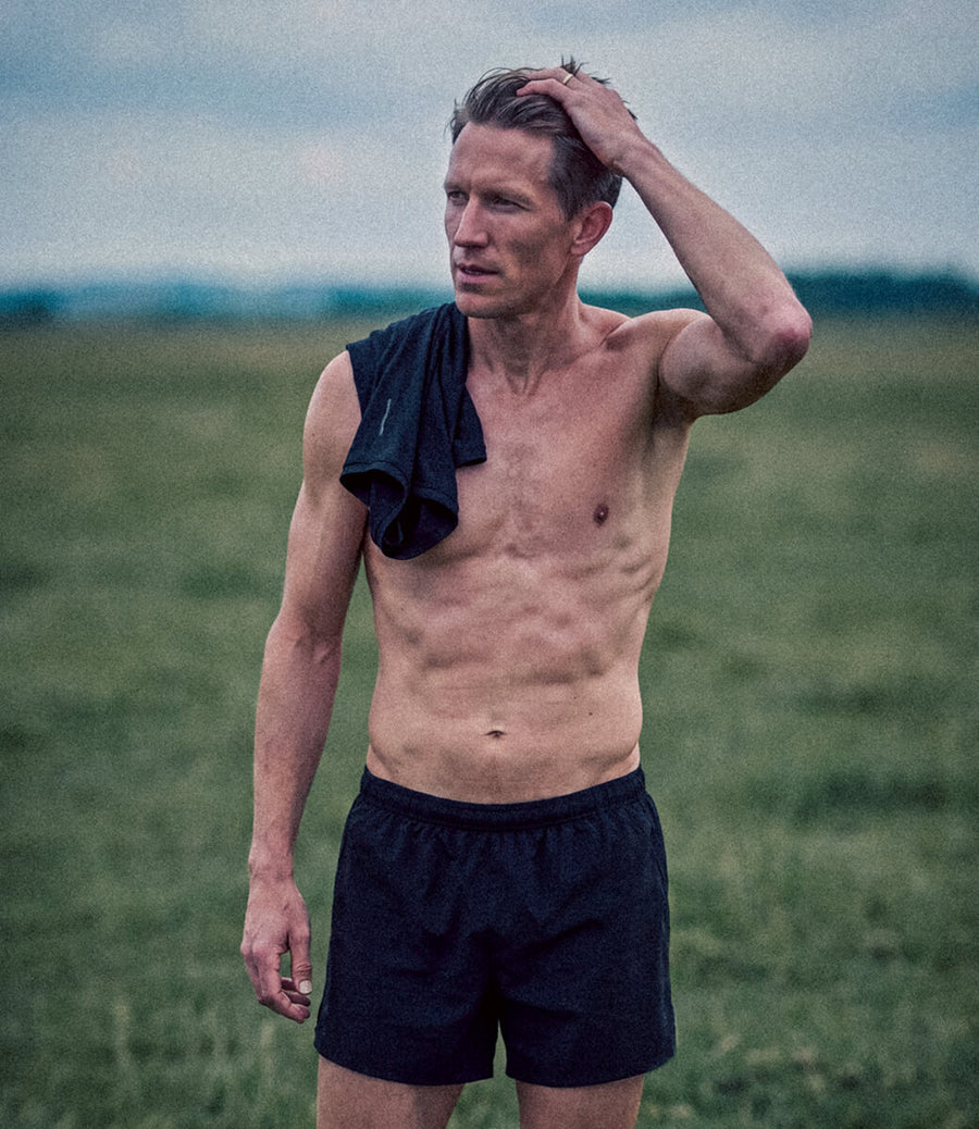 A shirtless man standing in an open grassy field, wearing dark athletic shorts and having a dark towel draped over one shoulder, with his hand running through his hair.