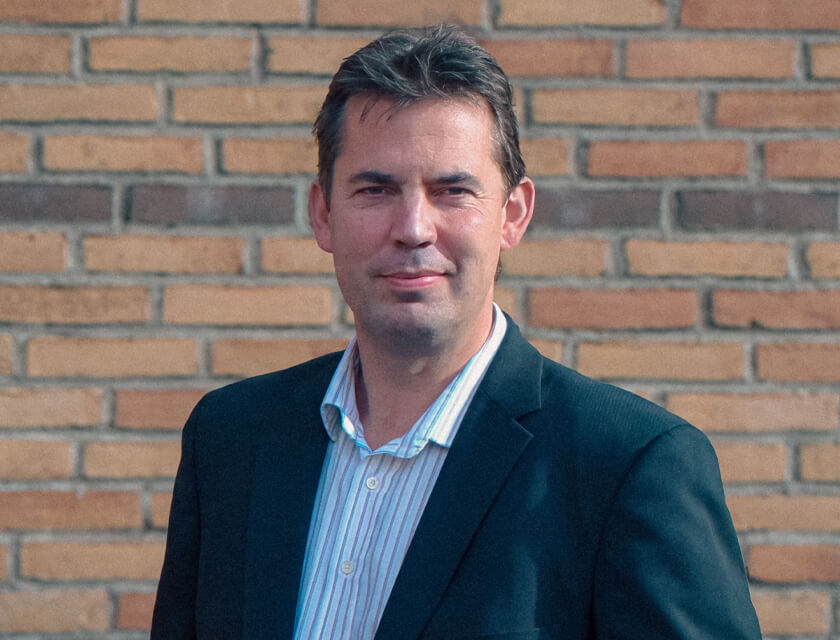 A man wearing a dark blazer and a light blue striped button-down shirt, standing in front of a brick wall.