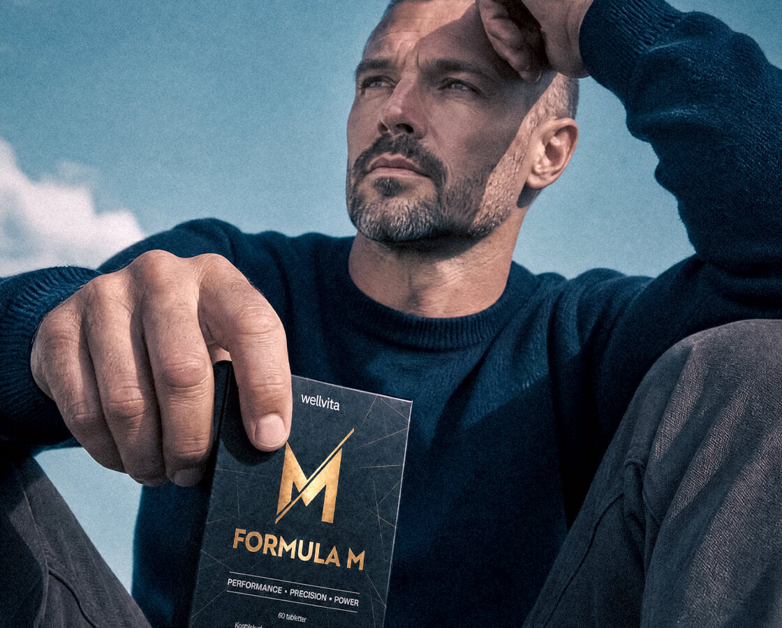 A man in a dark navy sweater holds a box of wellvita Formula M outdoors against a blue sky background.