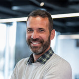 A smiling man with a beard and short hair, wearing a light gray crewneck sweater over a green plaid button-down shirt, photographed in a modern office setting.