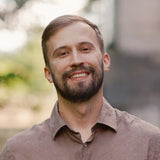 A smiling man with a beard wearing a brown button-up shirt, photographed outdoors with a softly blurred natural background.